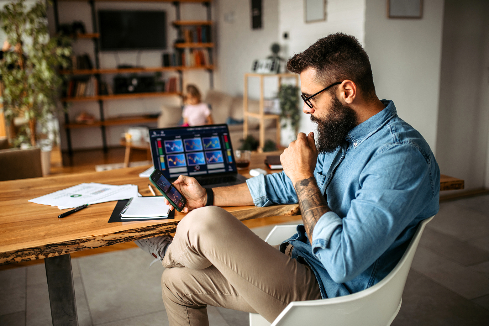 A man with a beard sits in front of a computer showing lots of graphs, looking at his phone thoughtfully.