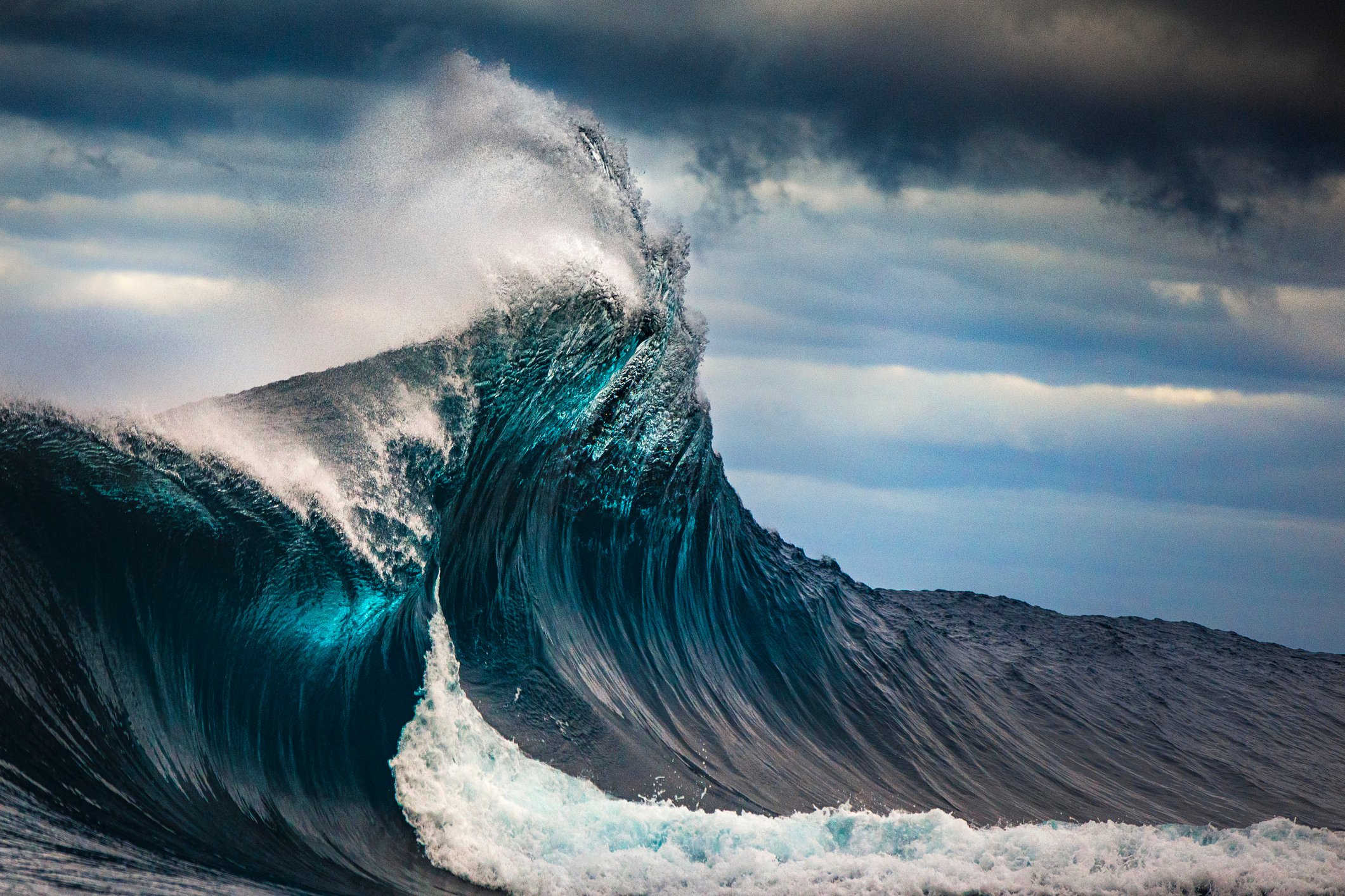 A large wave curls in front of a blue sky with white clouds